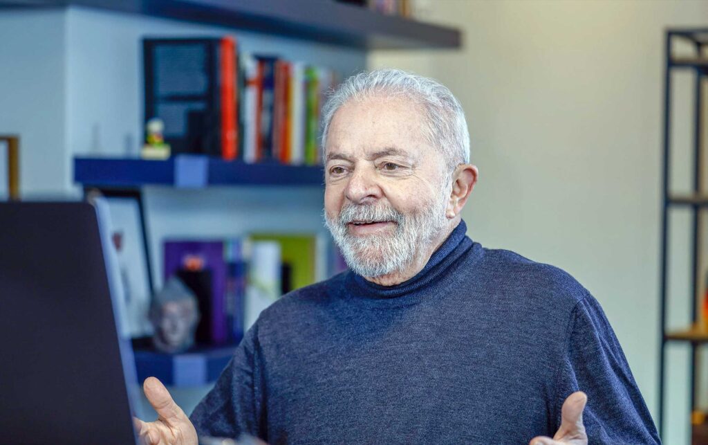 Presidente Lula, de braços abertos, sorri diante da tela de um computador